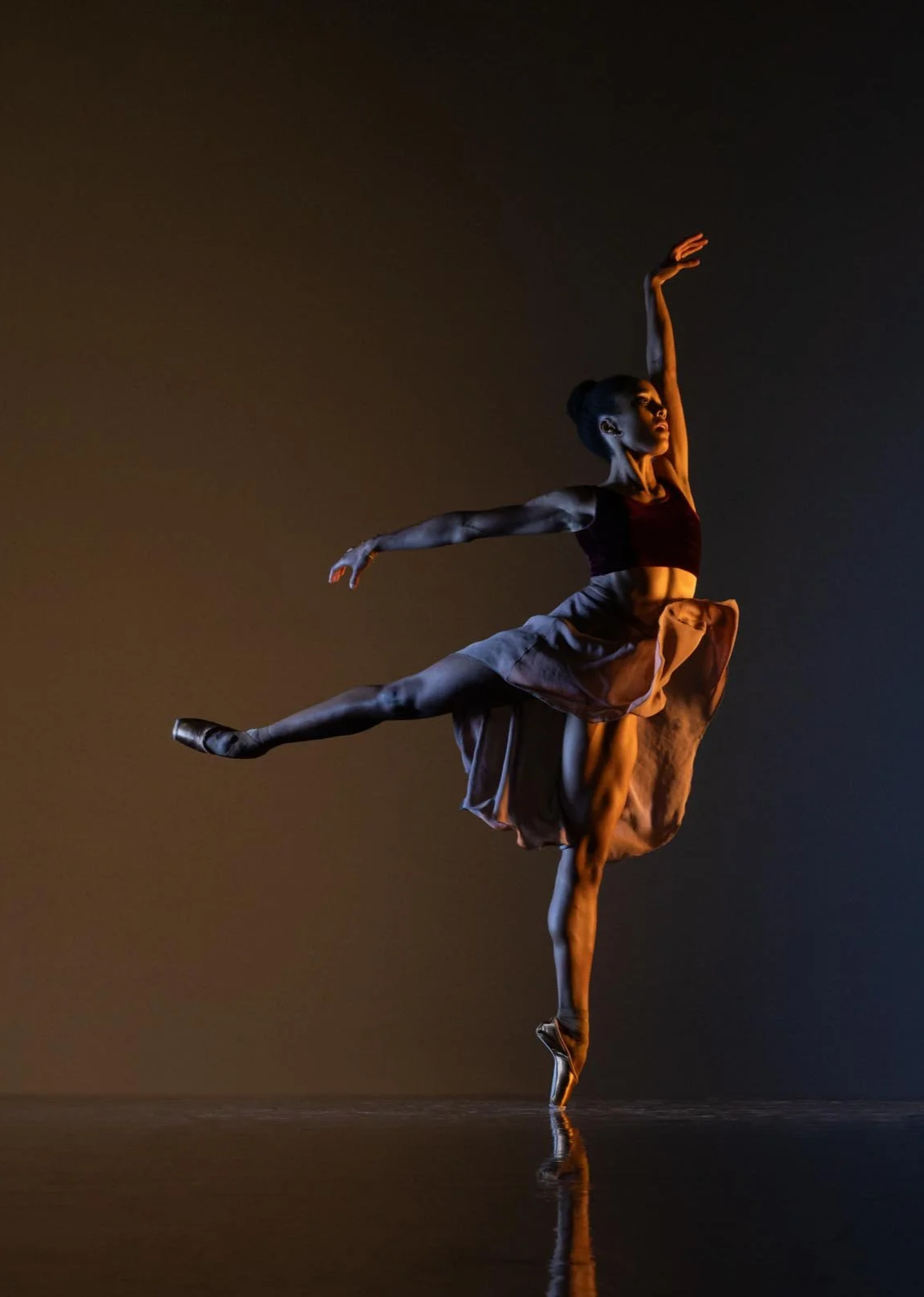 Ballet dancer performing a pose on pointe with one leg extended, arms in graceful positions, wearing a dark top and flowing skirt, against a dark background.