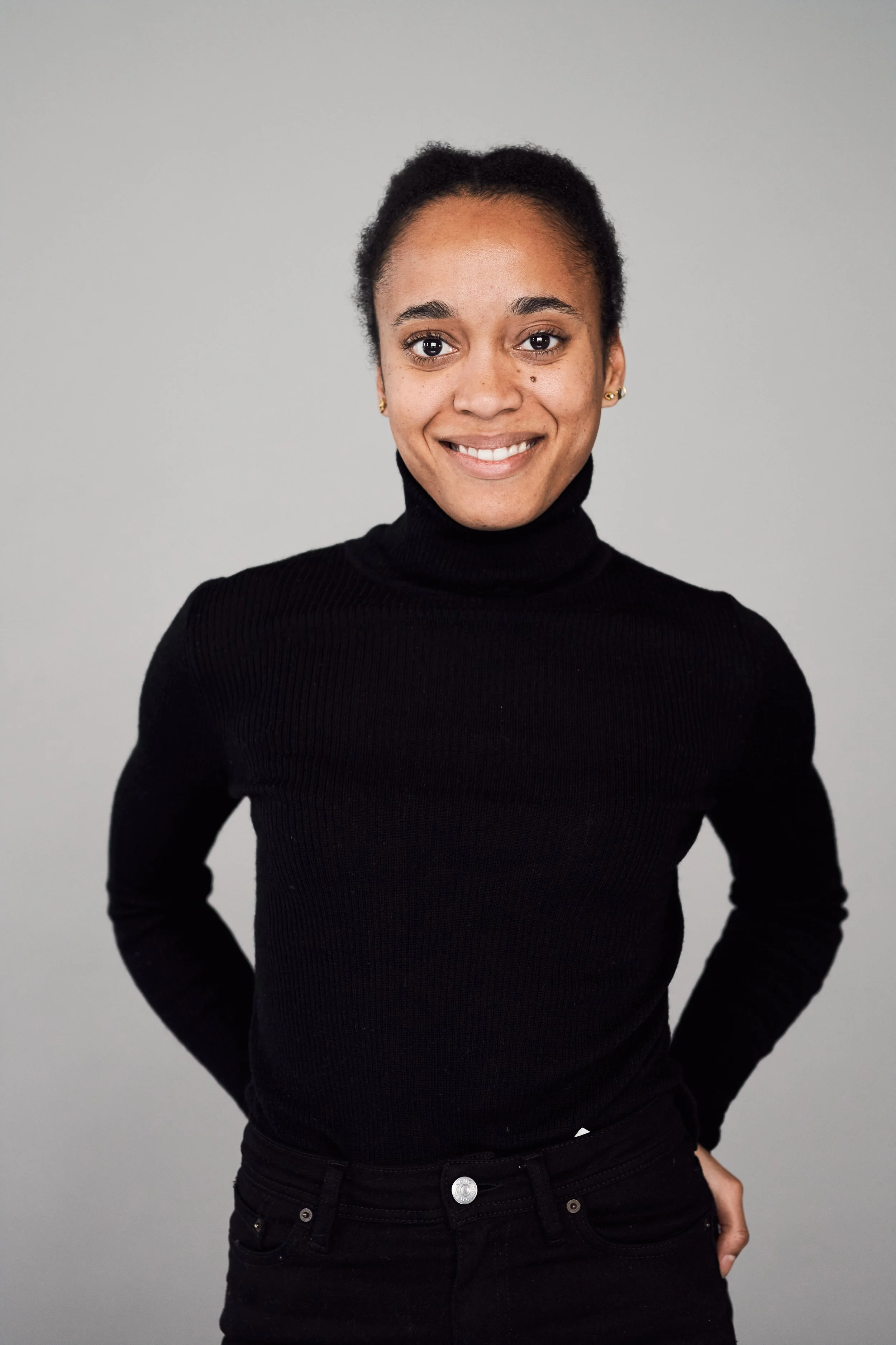 A young woman with dark hair pulled back, wearing a black turtleneck sweater and black pants, standing against a plain light gray background, smiling at the camera.