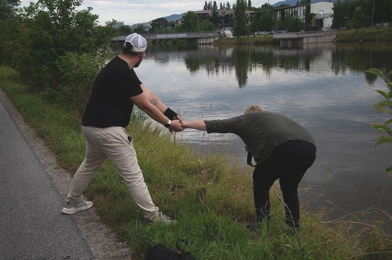 Zwei Menschen am Flussufer, einer zieht den anderen aus dem Wasser, während er sich nach vorne beugt.