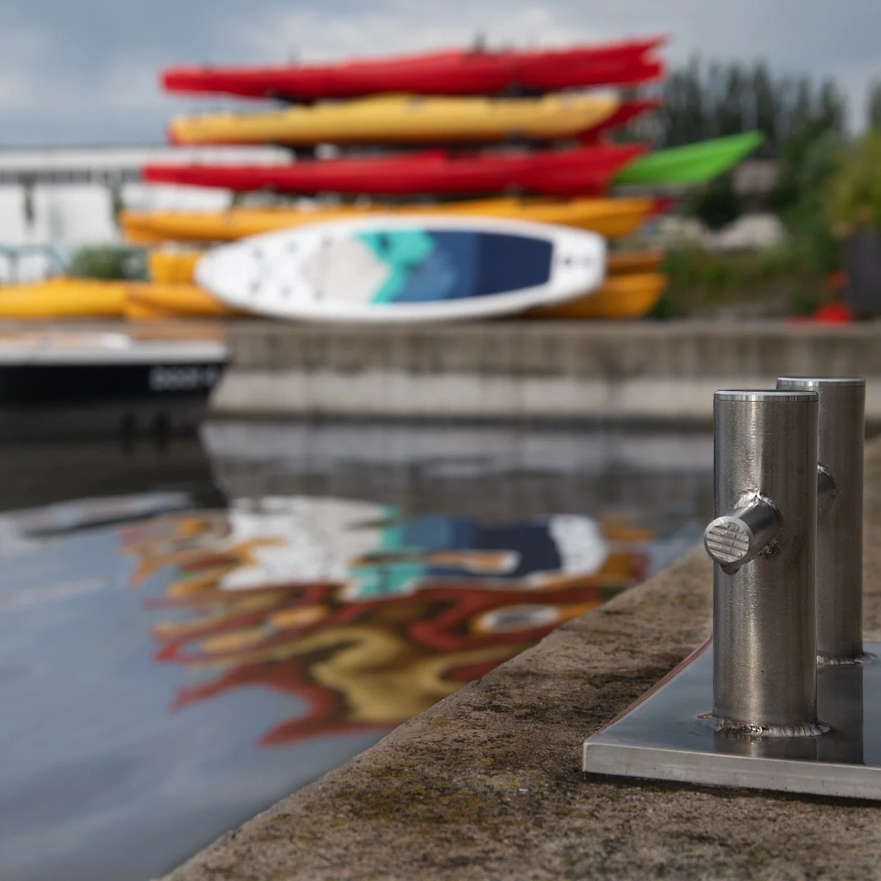 Blick auf einen Bootssteg mit mehreren aufgestapelten bunten Kanu- und Stand-up-Paddle-Boards im Hintergrund, während im Vordergrund ein Edelstahlpfosten mit Gelenk und Schraube am Steinfuß sichtbar ist.