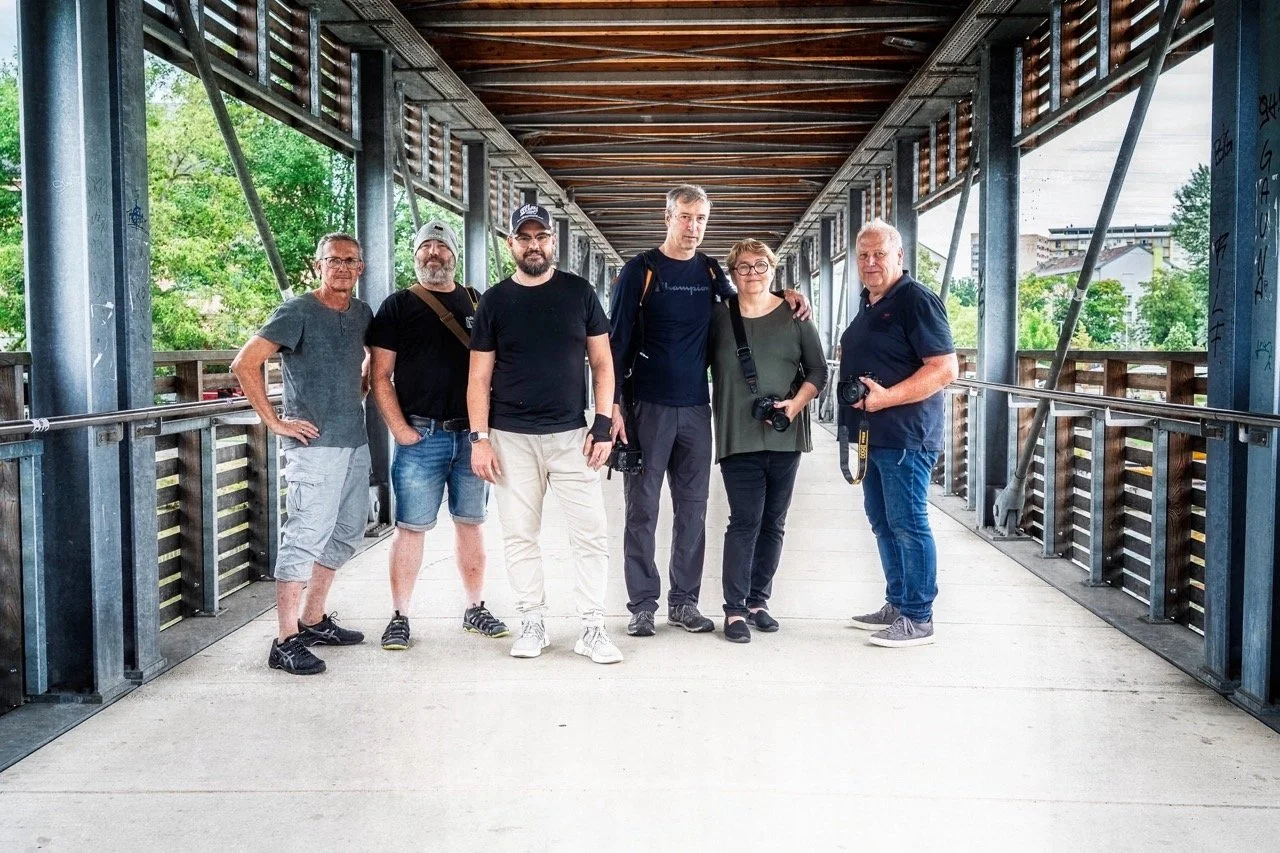 Sechs Menschen stehen auf einer Brücke, einige halten Kameras, die Umgebung ist grün und urban.