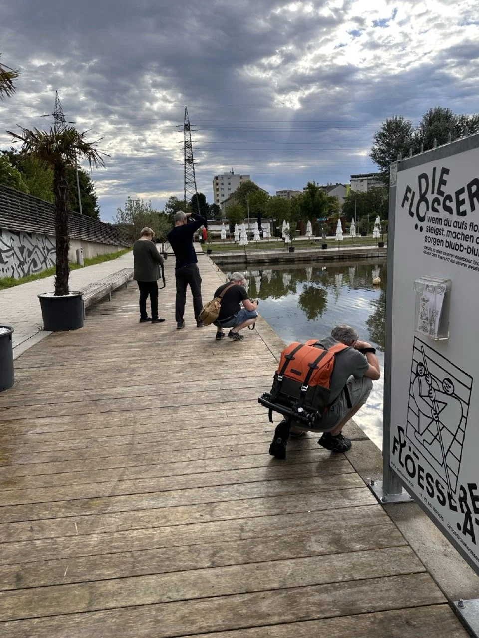 Vier Menschen beim Beobachten eines Teiches auf einer Holzpromenade, im Hintergrund Bäume, Gebäude und dunkler Himmel mit Wolken, rechts ein Infostand zur Flosser.