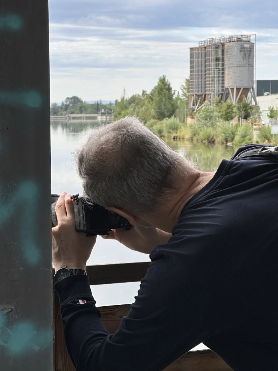 Mann fotografiert mit einer Kamera auf einer Brücke die Flusslandschaft mit Bäumen und einem Industriegebäude im Hintergrund.