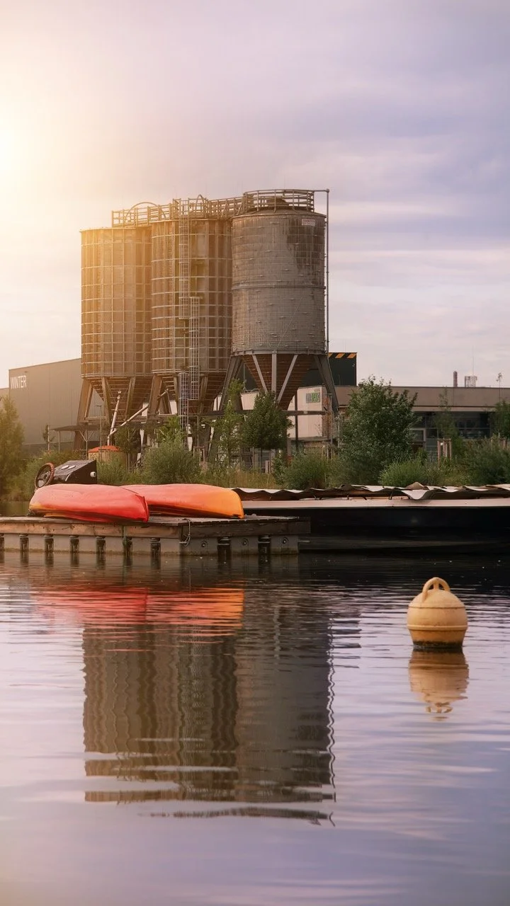 Silhouette alter Fabriksilos in der Dämmerung, neben einem Wasserweg mit aufgelegten Booten und einer Boje.