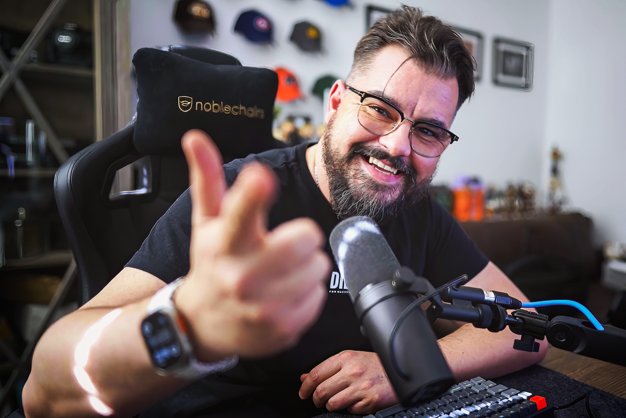 Mann mit Brille und Bart, lachend, in einem Gaming-Stuhl sitzend, winkt zur Kamera, im Hintergrund sind Helme an der Wand.