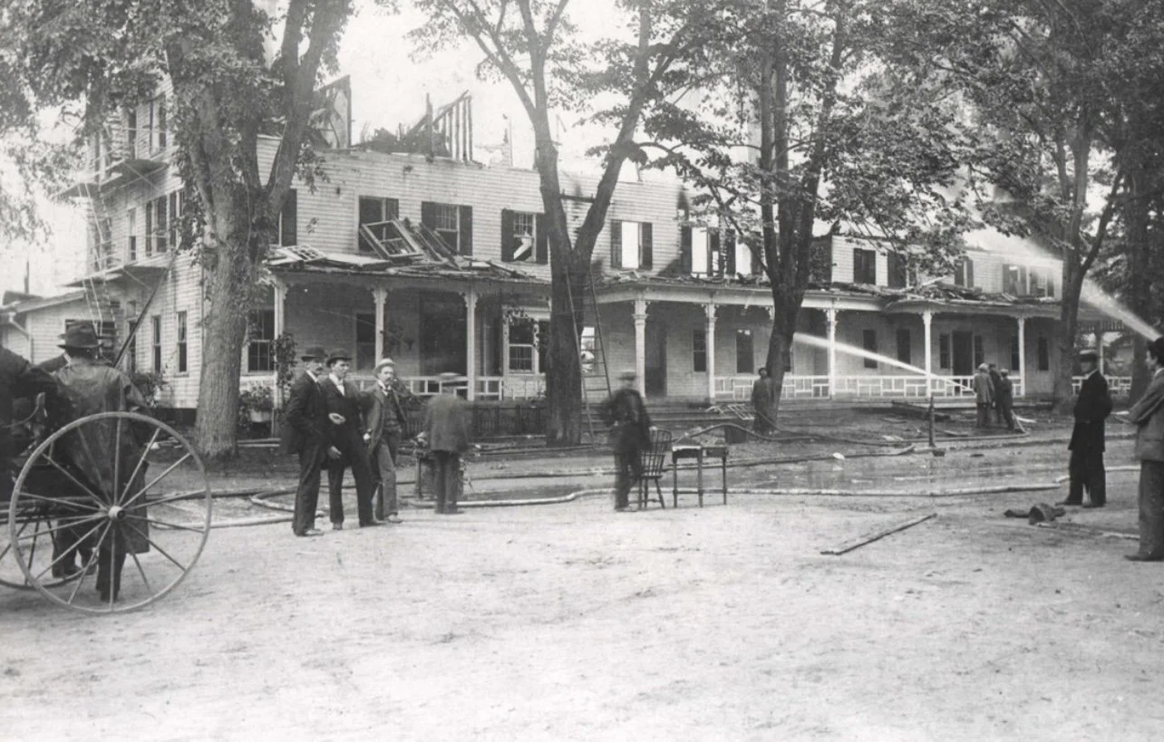 A black and white photo of a house on fire with firefighters spraying water from hoses. Several people are standing outside, some watching and others involved in firefighting efforts. There is a large tree and old-fashioned fire equipment in the scene.