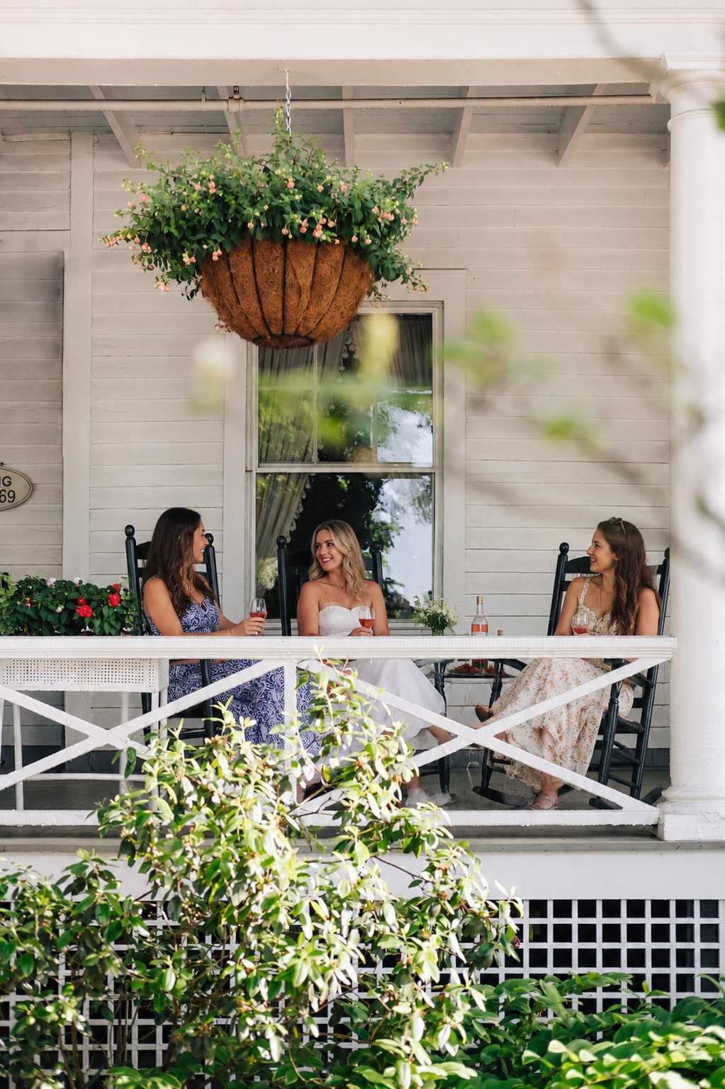 Three women sitting on a porch with black rocking chairs, having drinks and chatting, with green leafy plants in the foreground and a hanging flower basket above, behind a white wooden house exterior.