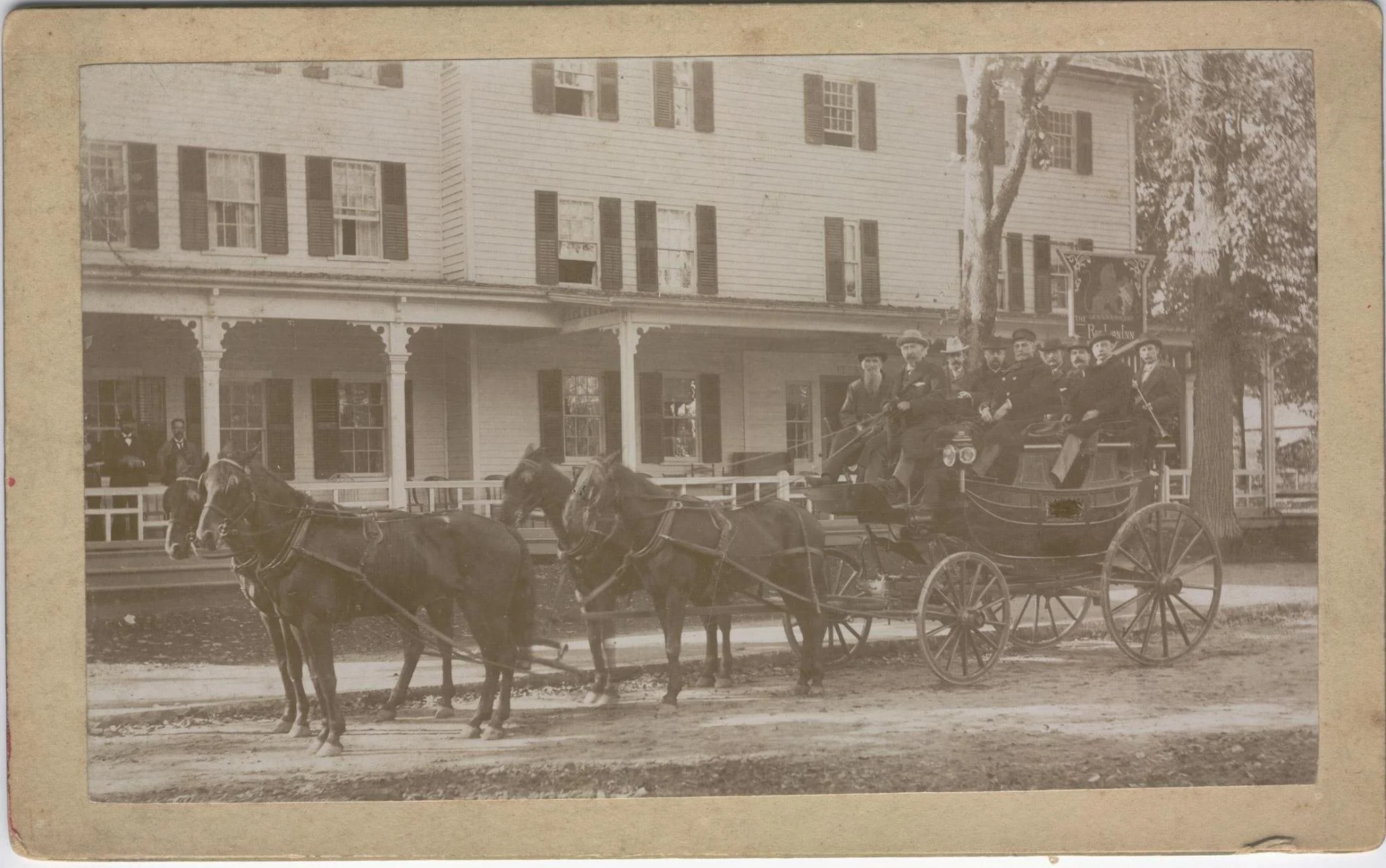 A vintage black and white photo showing a horse-drawn carriage with people seated inside, parked in front of a large, wooden, multi-story building with multiple windows and shutters, and a porch with decorative columns. There are a few men standing around and in the background, there are trees and signs.