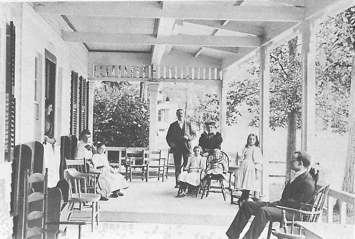 Black and white photograph of a group of people on a porch with trees in the background. There are children and adults sitting or standing, with some chairs lined up along the porch.