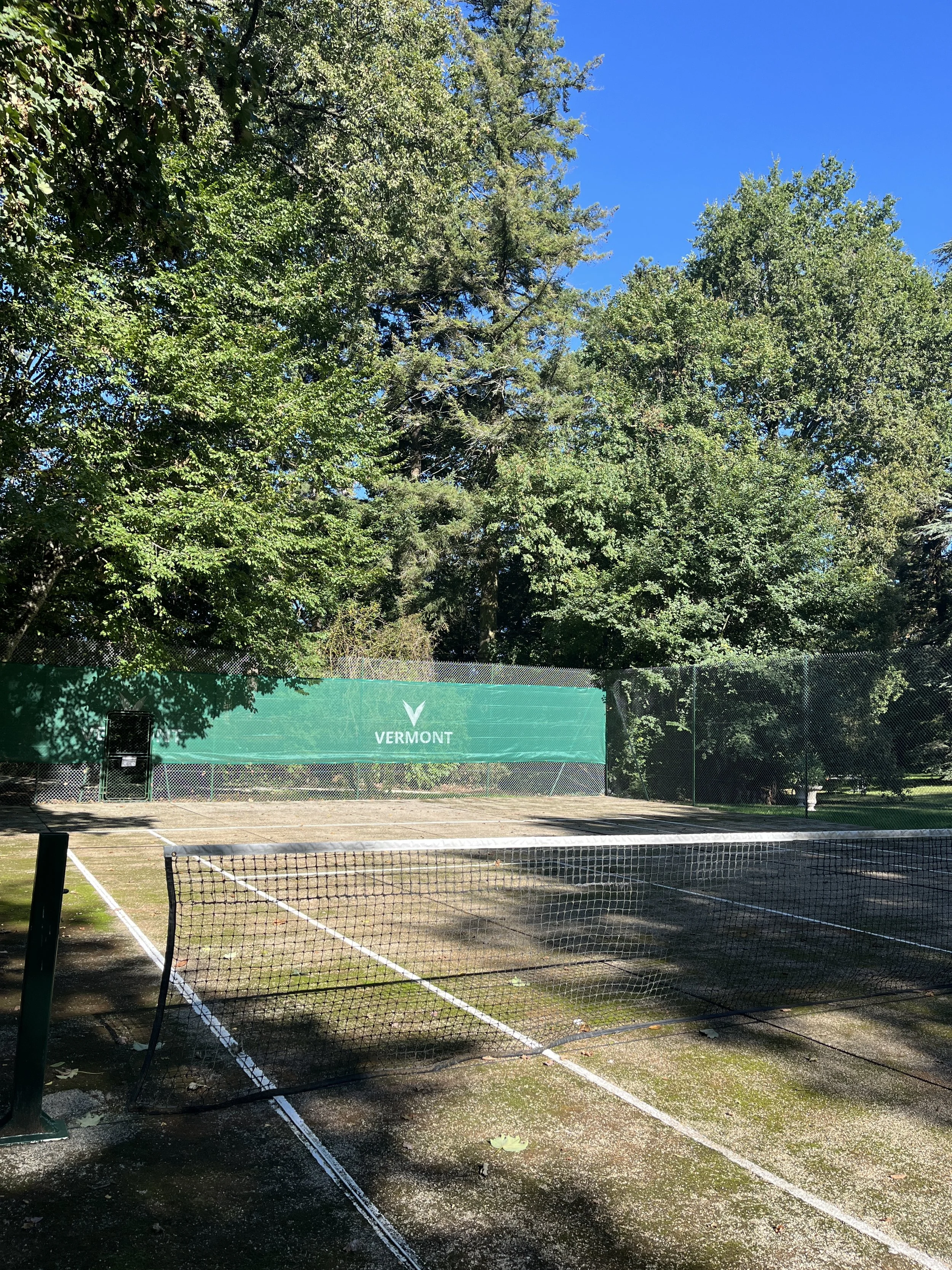 Empty outdoor tennis court surrounded by trees with green leaves; green banner with the word 'Vermont' on the back wall; clear blue sky.
