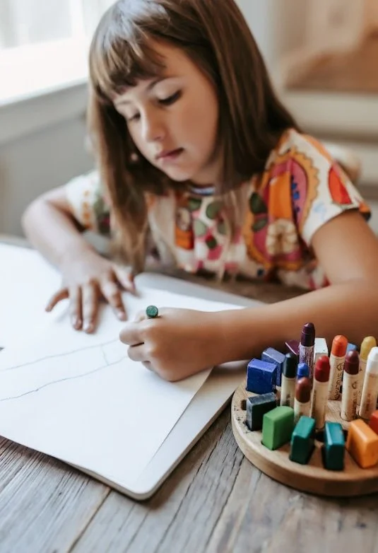 A young girl with brown hair drawing on a paper with a green marker, sitting at a wooden table near a window, with a round tray of colorful markers nearby.