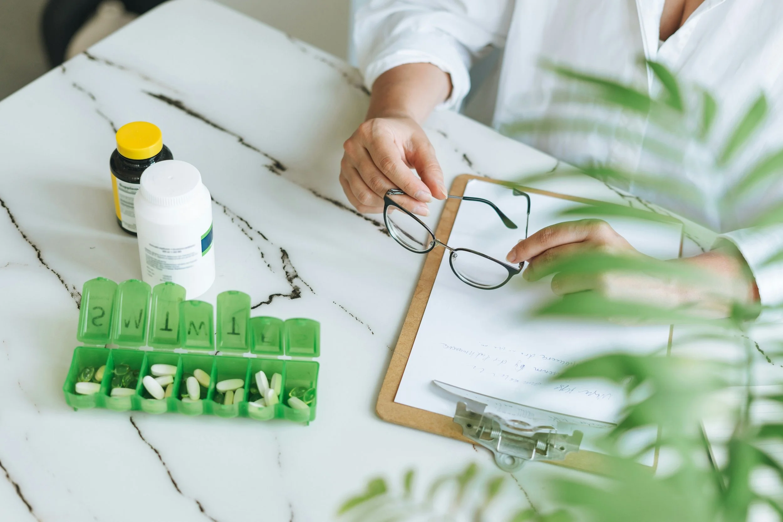 A person sits at a white marble table with a few bottles of pills, a green pill organizer, and a clipboard with handwritten notes. The person is holding a pair of glasses in one hand.