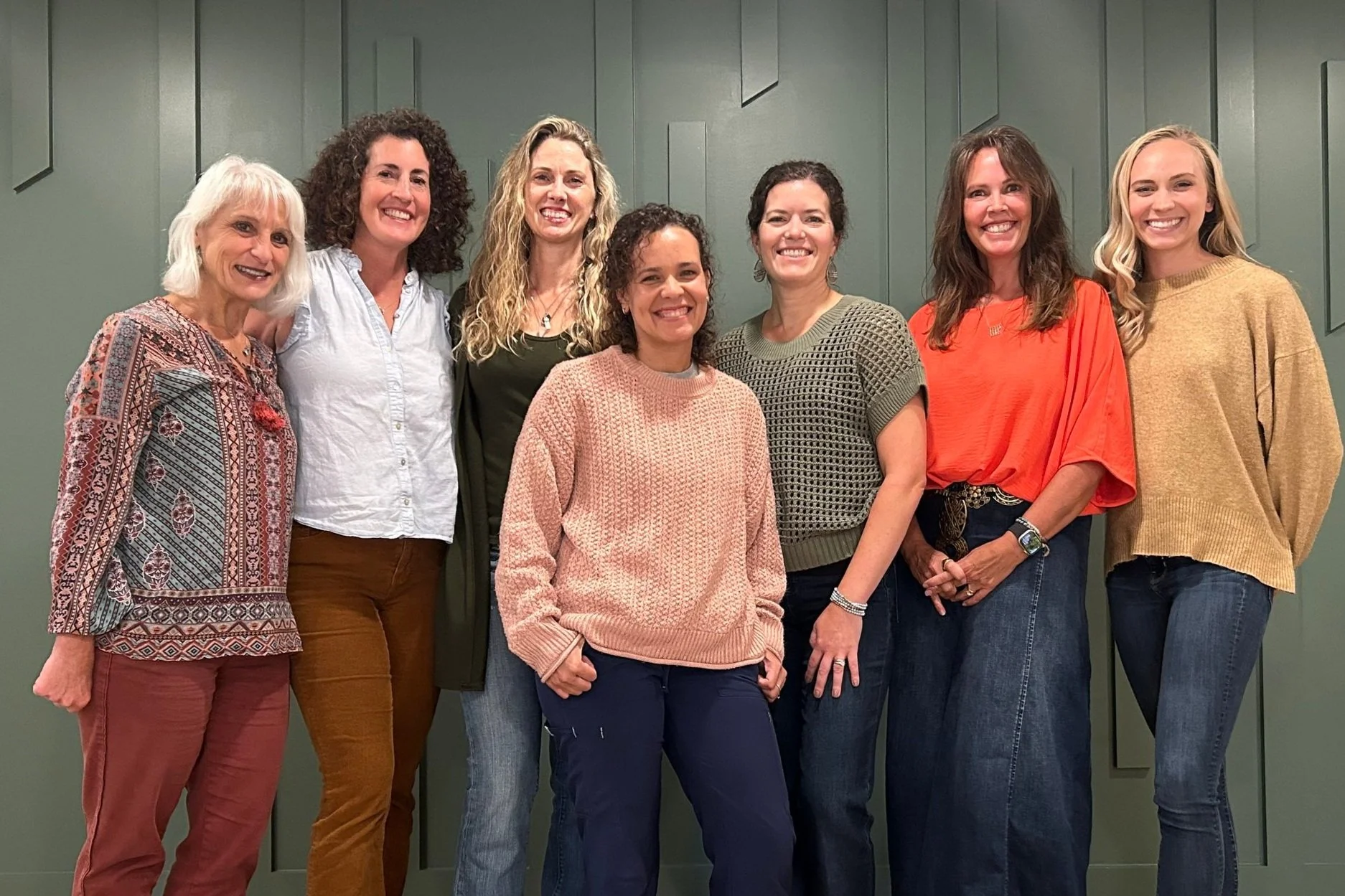 Group of eight women standing together indoors, smiling, against a green wall with paneling.