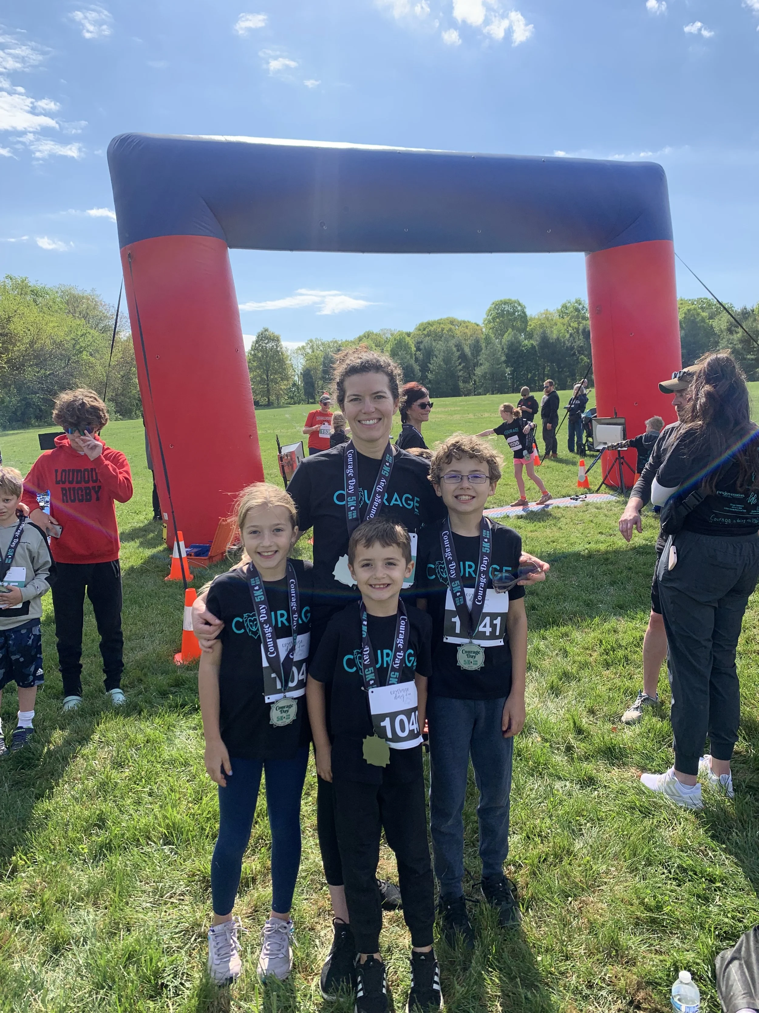 A woman and three children standing together smiling after finishing a race at an outdoor event, with a large inflatable arch in the background and other participants around.