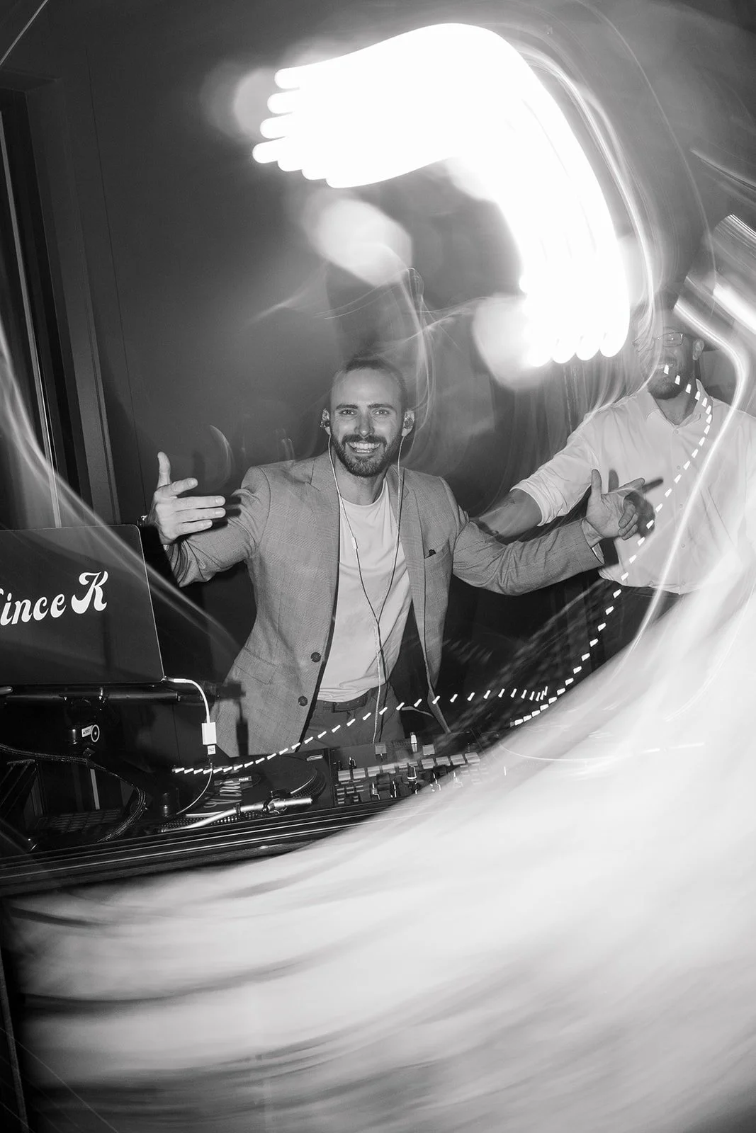 Vincent Kendjorsky  is DJing at a party with a DJ mixer and laptop. He is dancing and gesturing with his hands. The photo has light streaks and motion effects, and a person in the background gesturing as well. This is at Jorgensen Oak Grove.