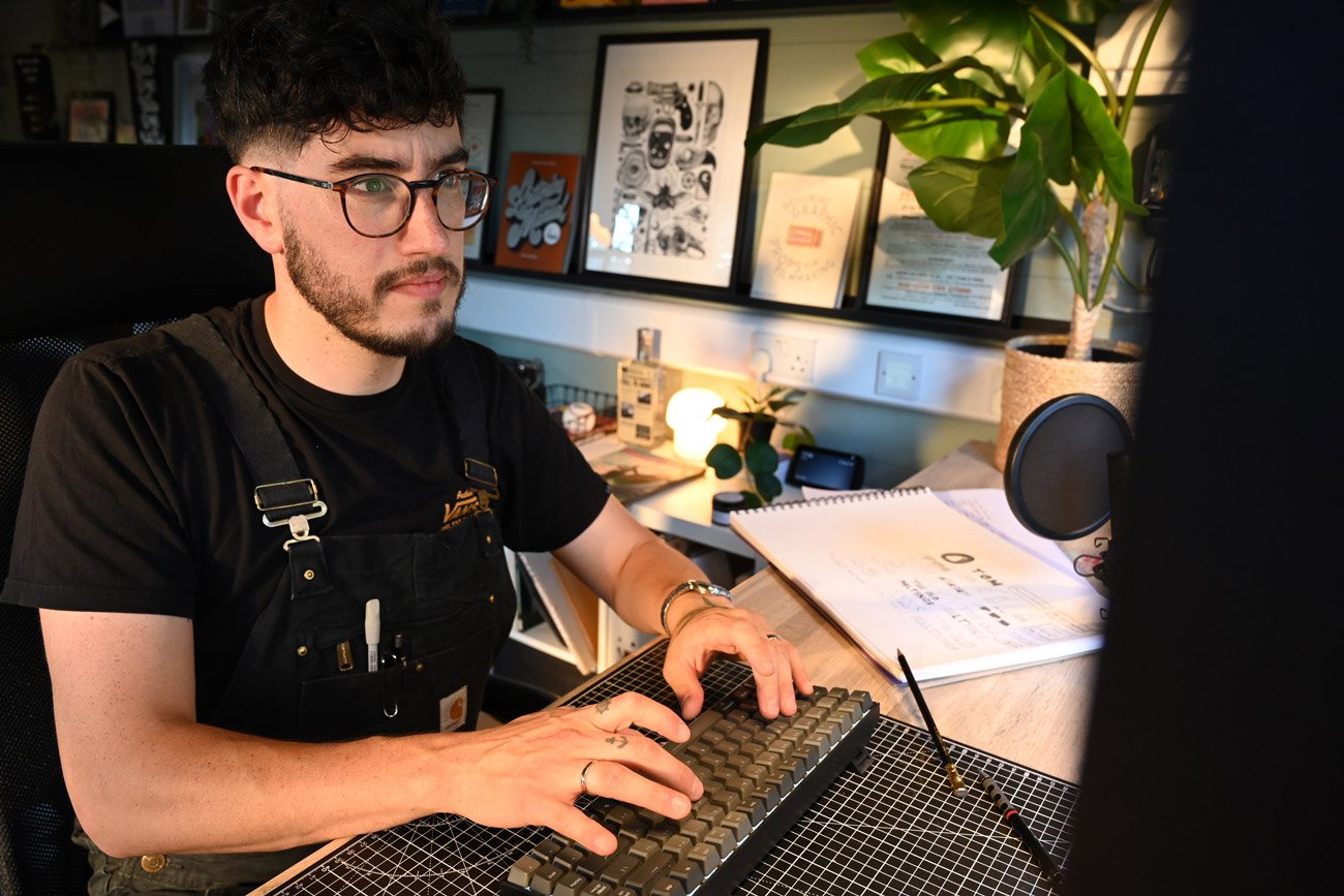 A man with glasses and a beard working on a computer in a cozy, decorated home office, surrounded by framed artwork, plants, and various office supplies.