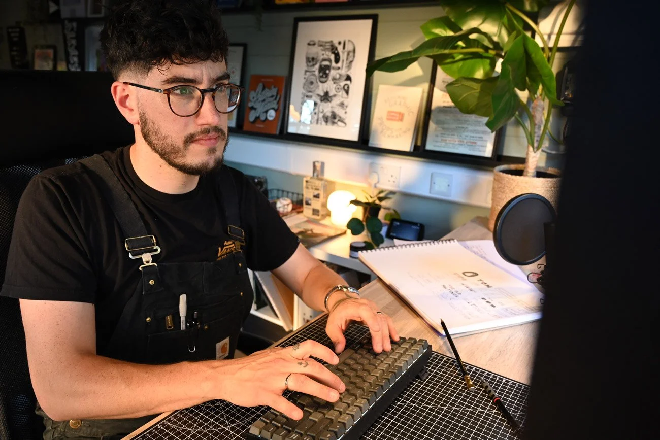 A man with glasses and a beard working on a computer in a cozy, decorated home office, surrounded by framed artwork, plants, and various office supplies.