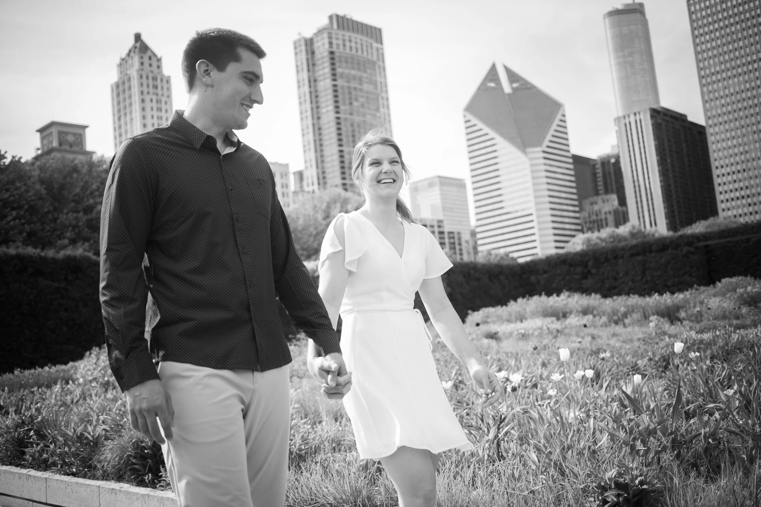 A couple walking hand in hand in a park with tall city buildings in the background.