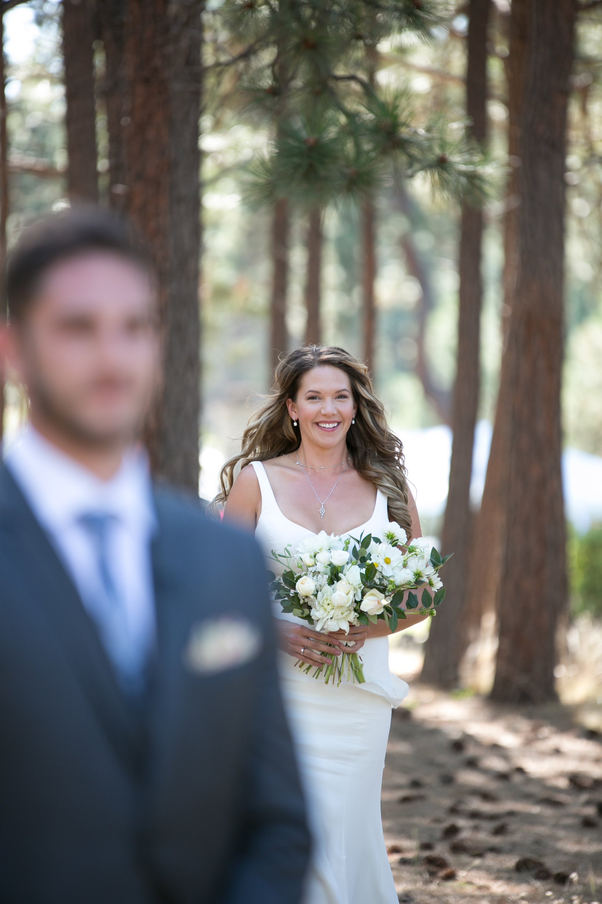 A bride in a white dress holding a bouquet approaches a man in a suit, outdoors among pine trees.