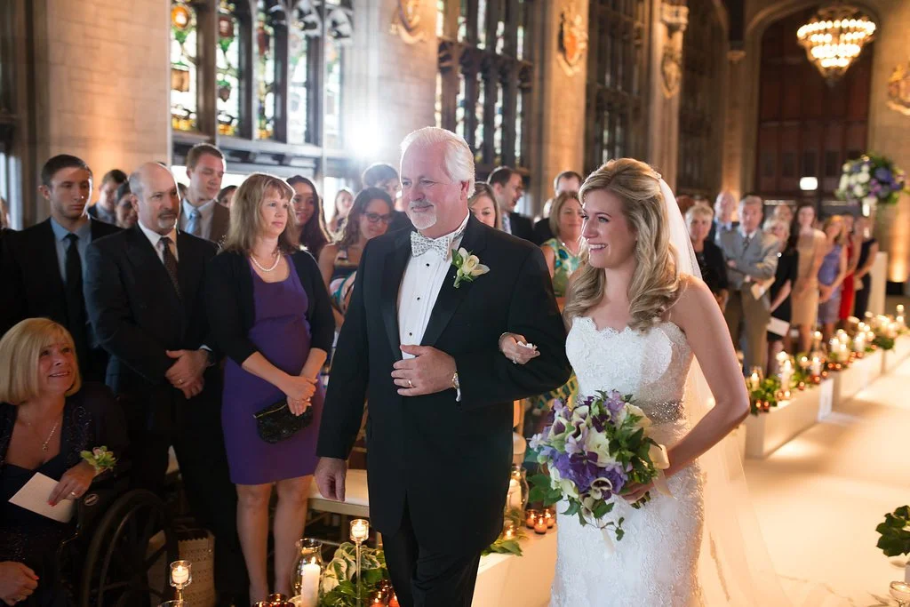 Bride walking down the aisle with an older man at a wedding ceremony, surrounded by guests in a decorated venue.