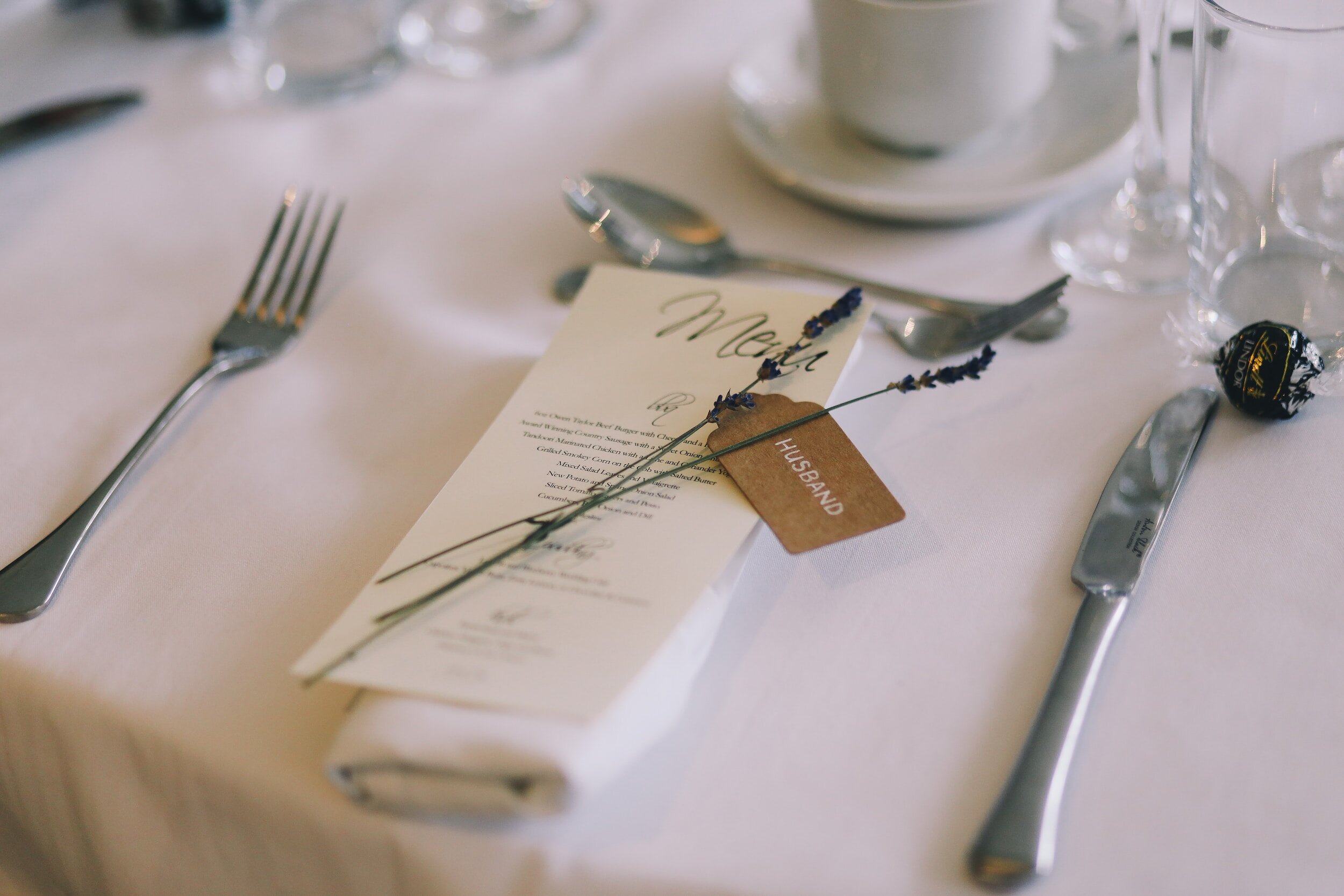 Table setting with fork, knife, napkin, menu, lavender sprigs, and a place card labeled "Husband" on a white tablecloth.