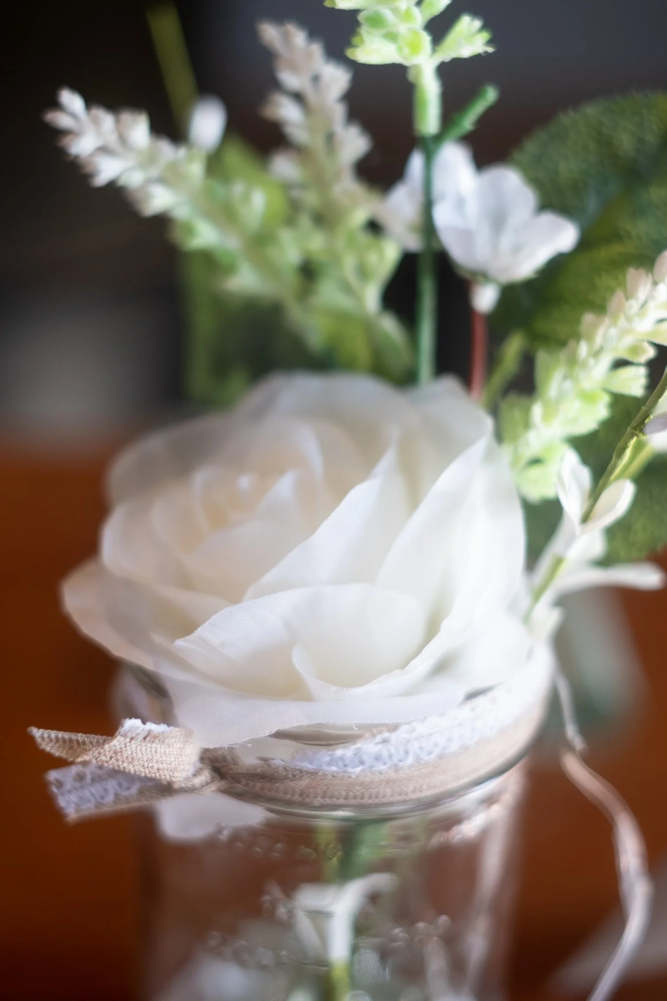 Close-up of a white artificial rose and green foliage in a glass jar, wrapped with lace and ribbon.