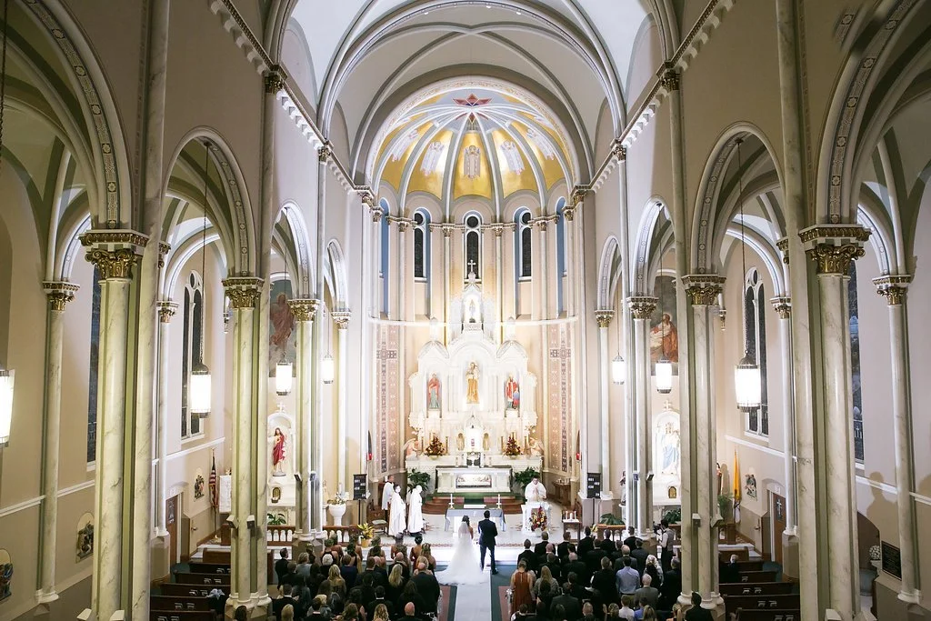 Interior of a large church with high arched ceilings and ornate columns, featuring a wedding ceremony taking place at the altar. The congregation is seated in wooden pews, and the church is decorated with statues and religious artwork.