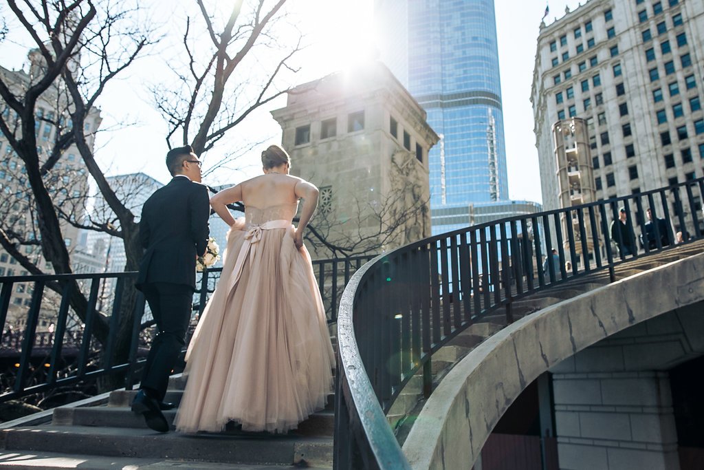 A couple dressed formally, with the woman in an elegant gown, walking up stairs outside near modern high-rise buildings.
