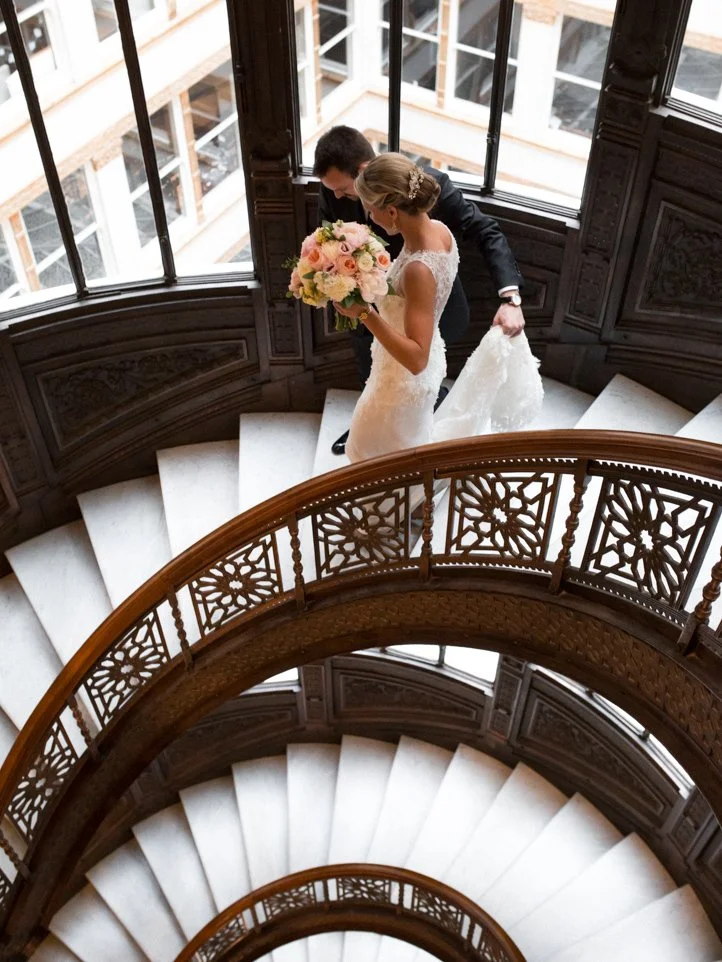 Bride and groom walking down a spiral staircase with ornate wooden railing, bride holding a bouquet of pink and white flowers.