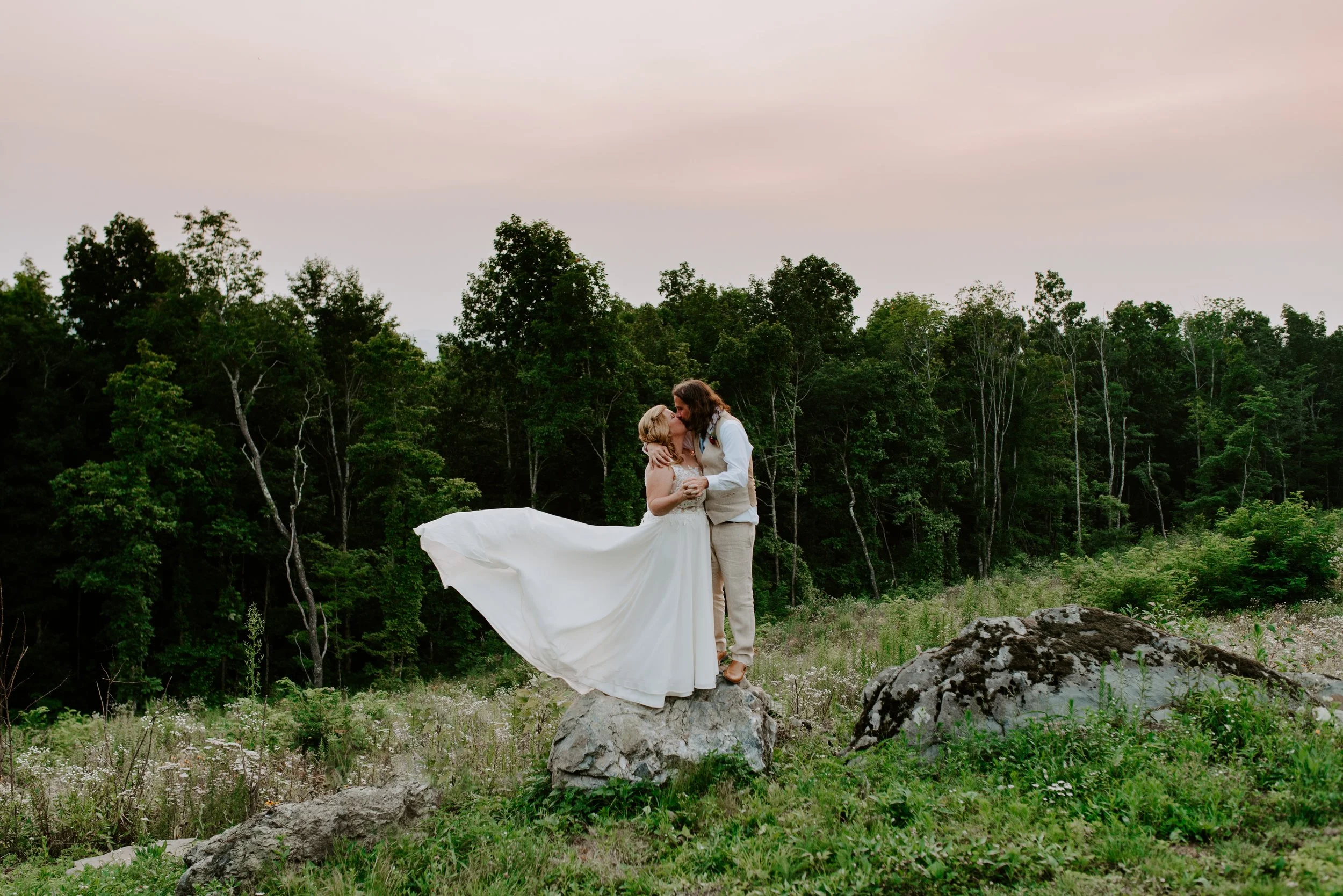 Bride and groom kissing on a rock in a forest setting, bride's dress flowing in the wind, with a backdrop of green trees and a pastel sky.