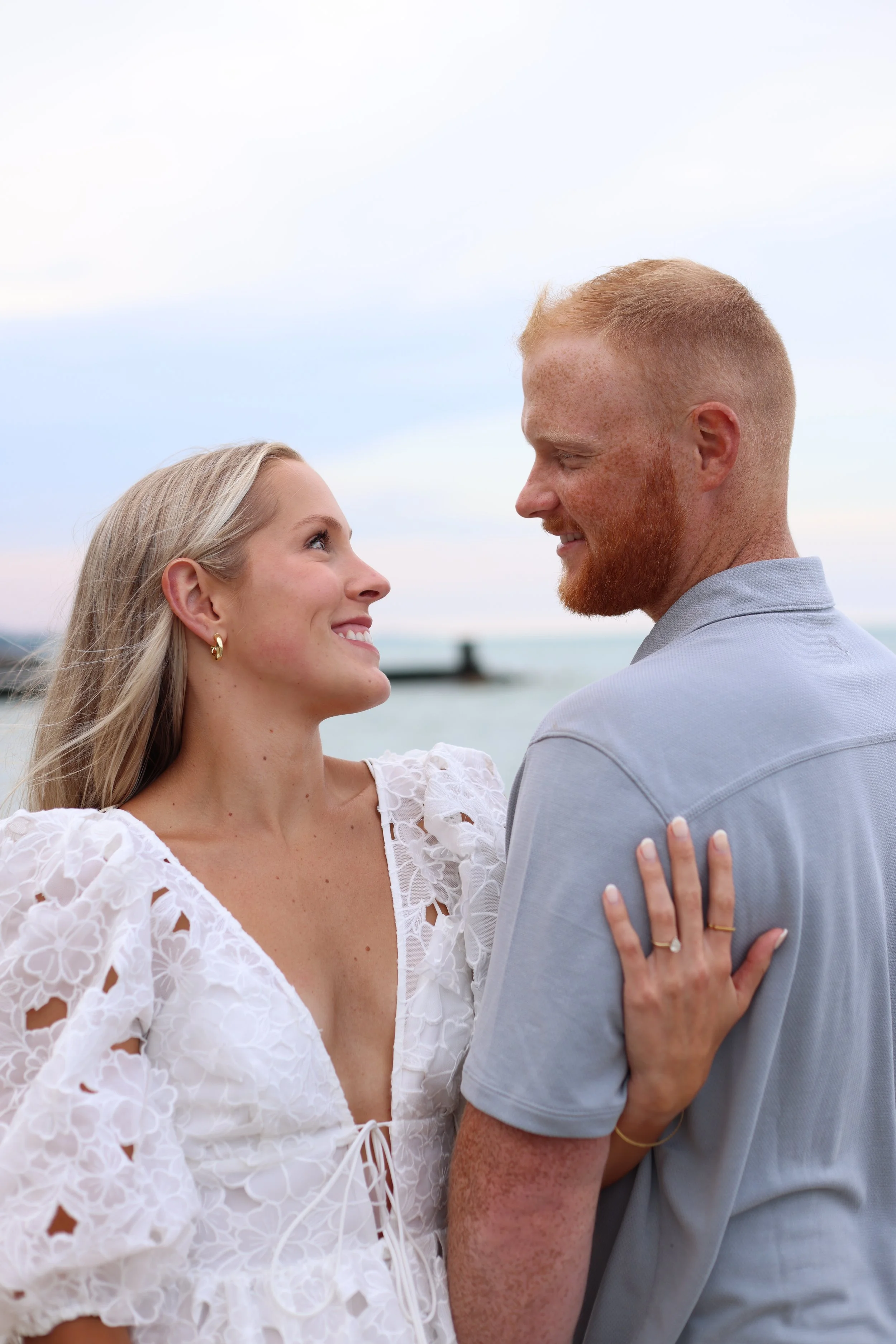 A couple smiling at each other by the sea, with the woman wearing a white floral dress and the man in a light blue shirt.