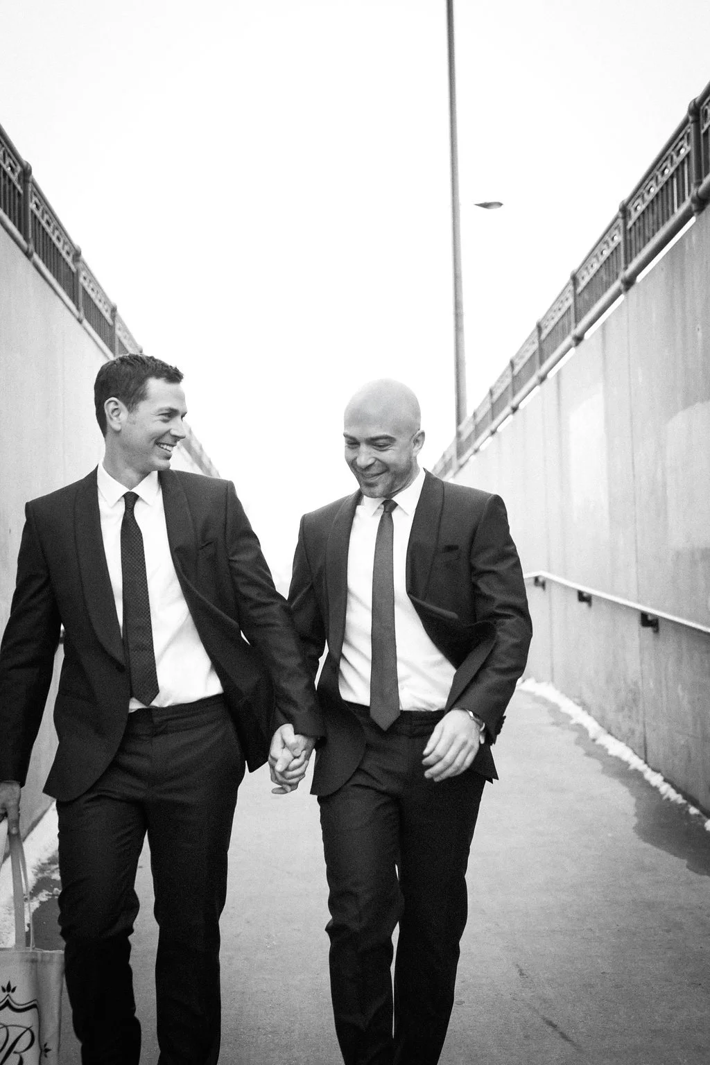 Two men in suits holding hands and smiling while walking down a pathway.
