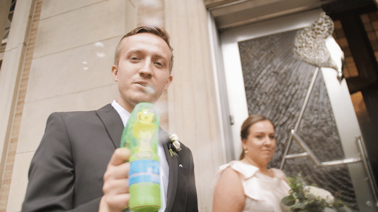 Man in suit blowing bubbles near woman in white dress, possibly at a wedding.