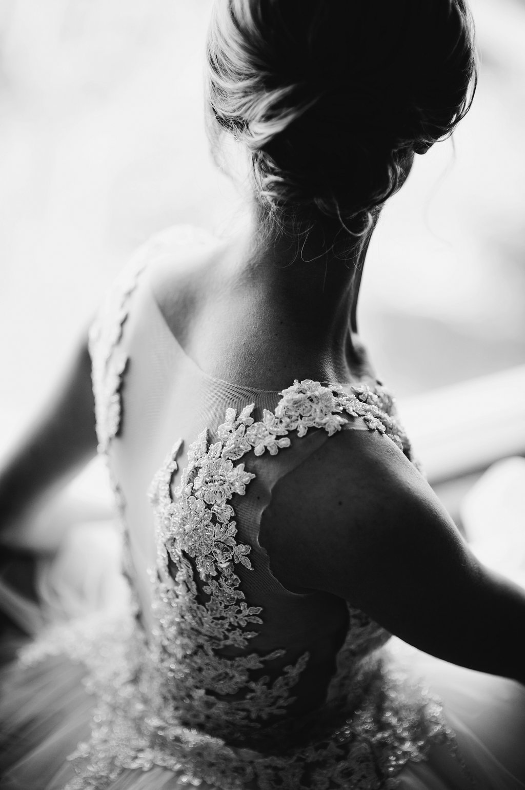 Black and white photo of a woman in an intricate lace dress, viewed from behind, with her hair in an elegant updo.