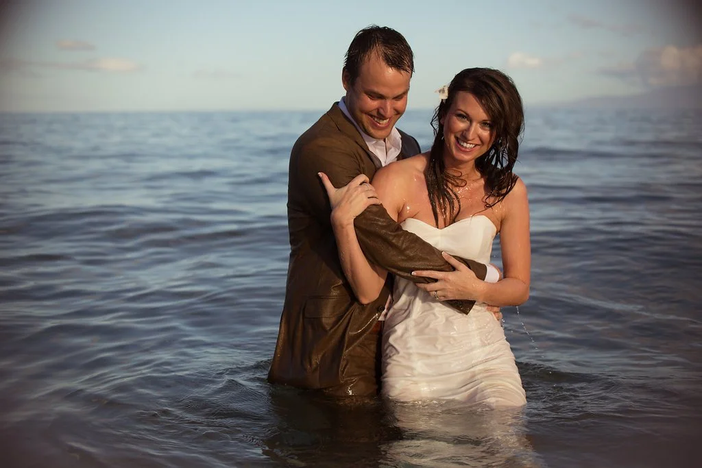 A couple in wedding attire standing waist-deep in the ocean, smiling and embracing.