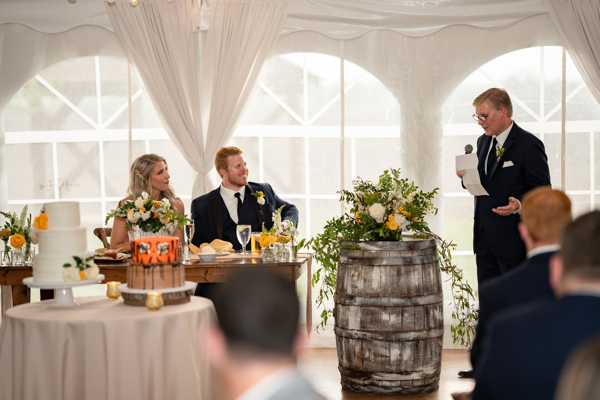 A wedding reception with a bride and groom seated at a table decorated with flowers, listening to a man in a suit giving a speech. A decorated barrel and cakes are displayed nearby.