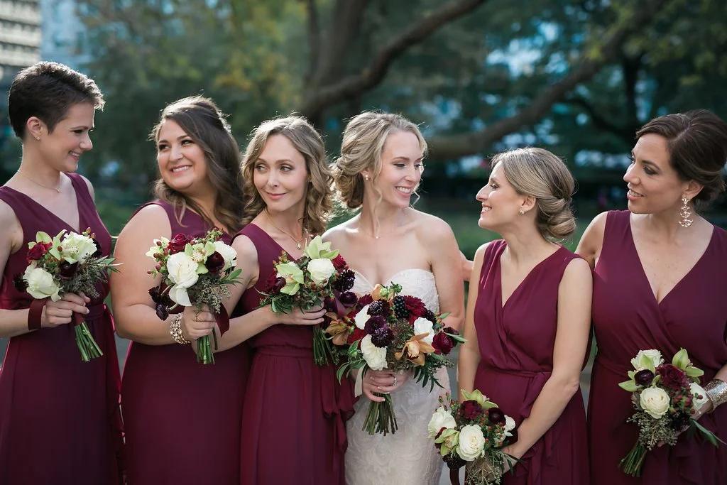 Bride with bridesmaids in dark red dresses holding bouquets, outdoors with trees in the background.
