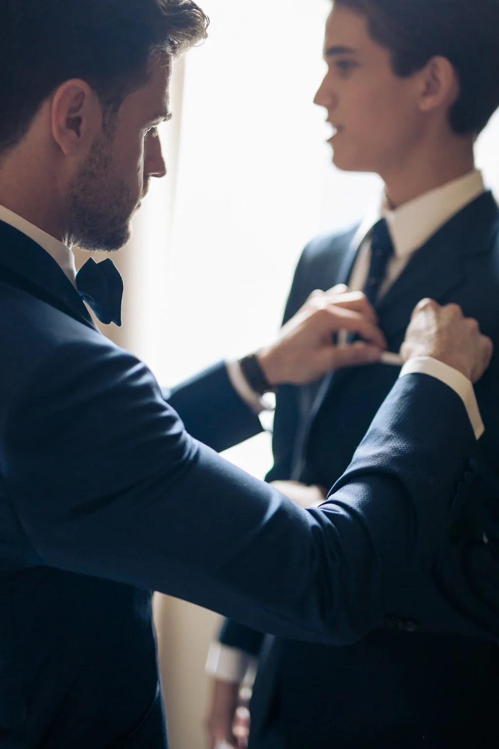 Two men in suits, one adjusting the other's tie, in a well-lit room.