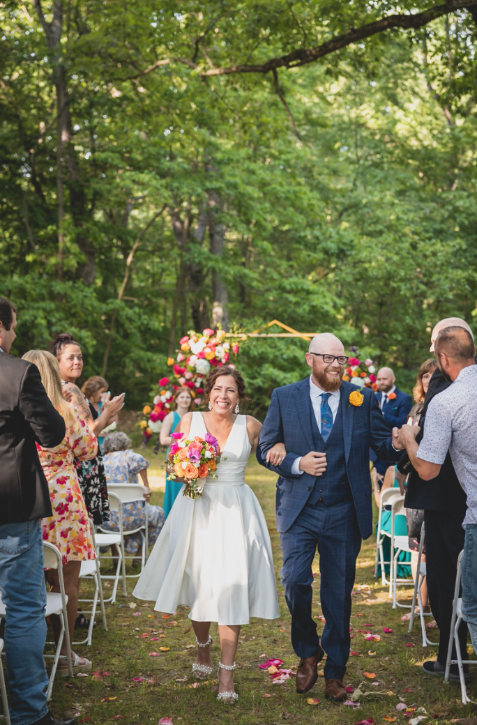 A newlywed couple joyfully walking down an outdoor aisle, the bride in a white dress holding a bouquet of colorful flowers and the groom in a blue suit. Guests are seated on either side, clapping and smiling. A floral arch is visible in the backgroun