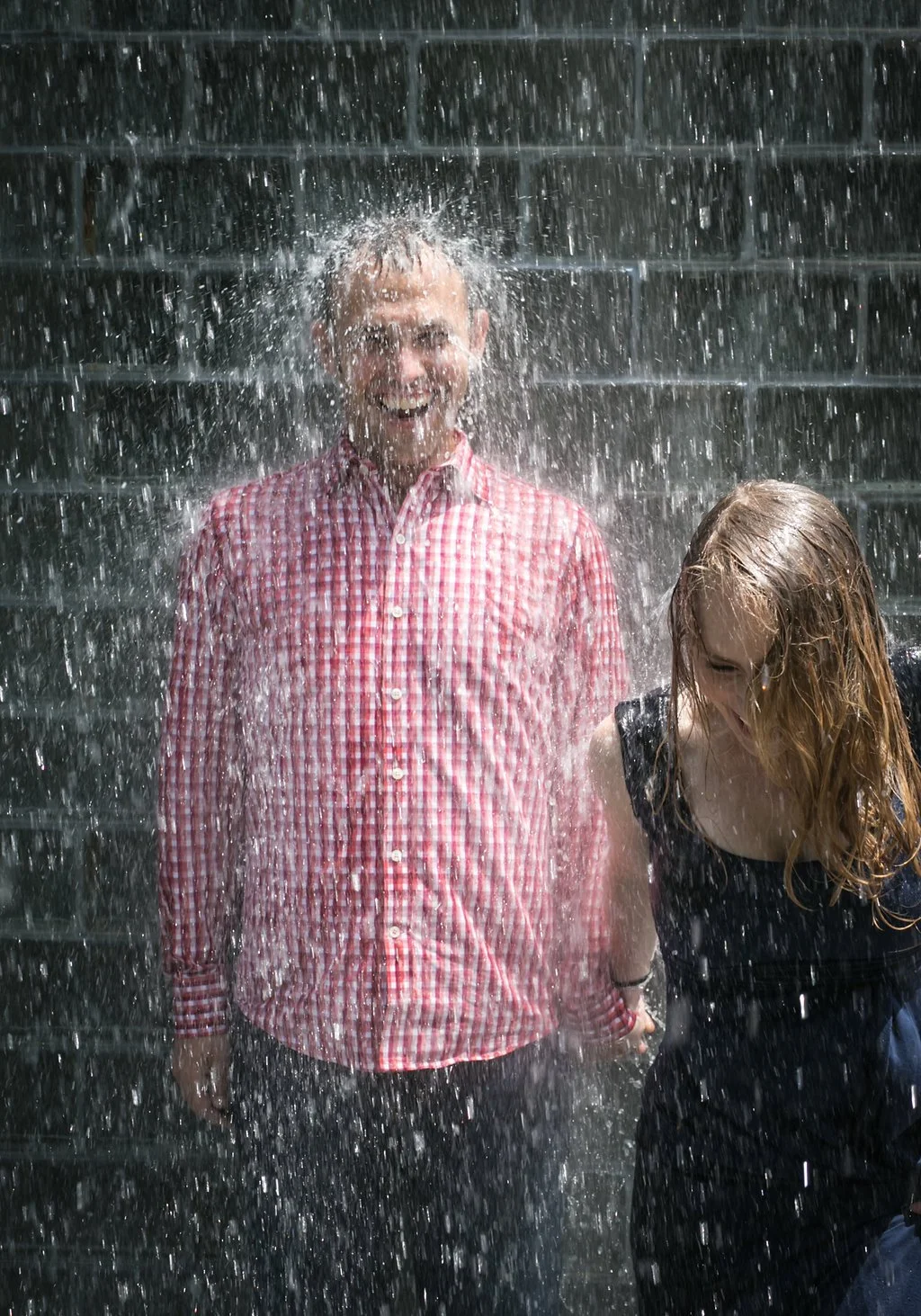 Two people standing under a waterfall, one in a red checkered shirt and the other in a black top, both smiling.