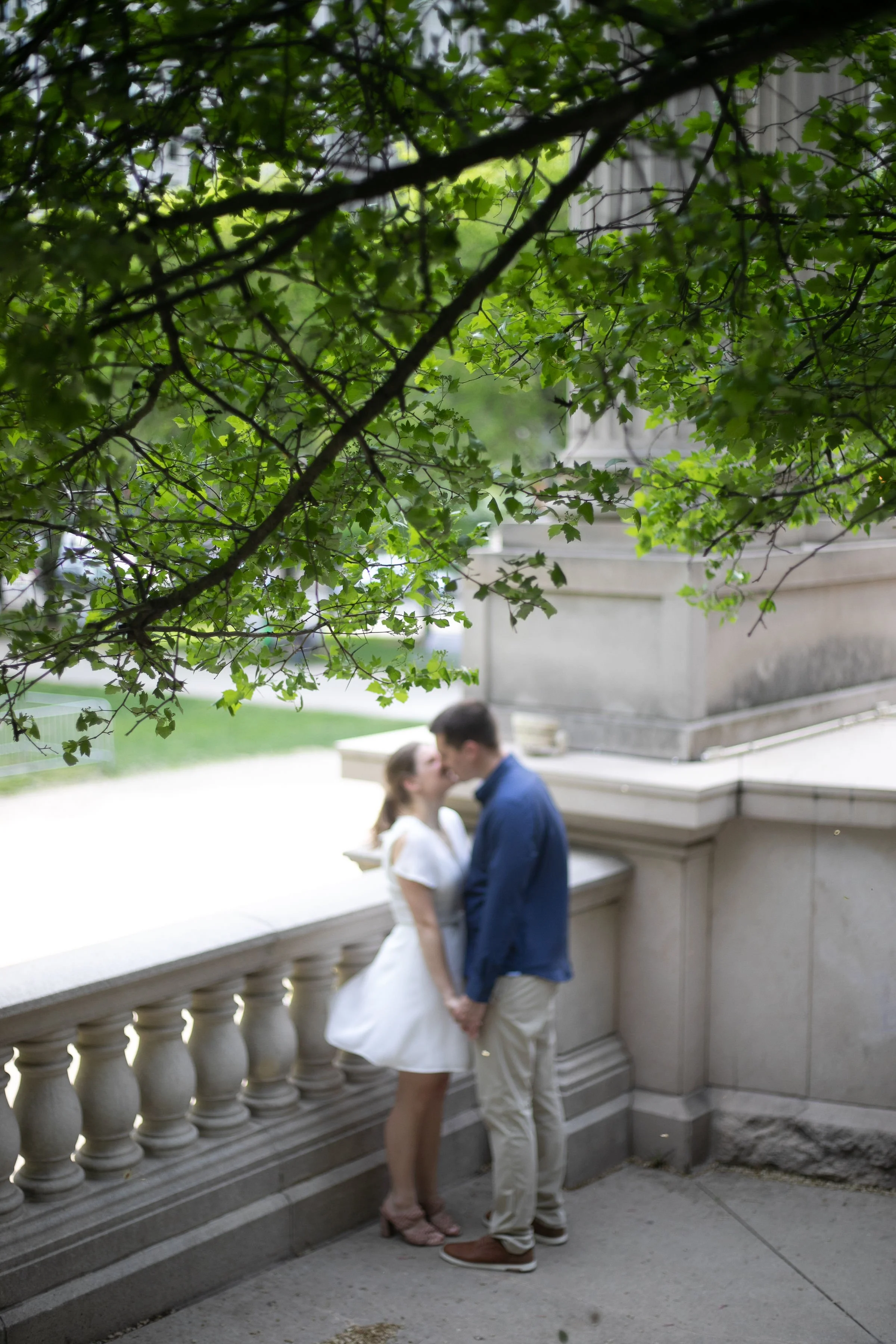 A couple standing on a stone terrace under a tree, holding hands and leaning towards each other, with blurred greenery in the background.