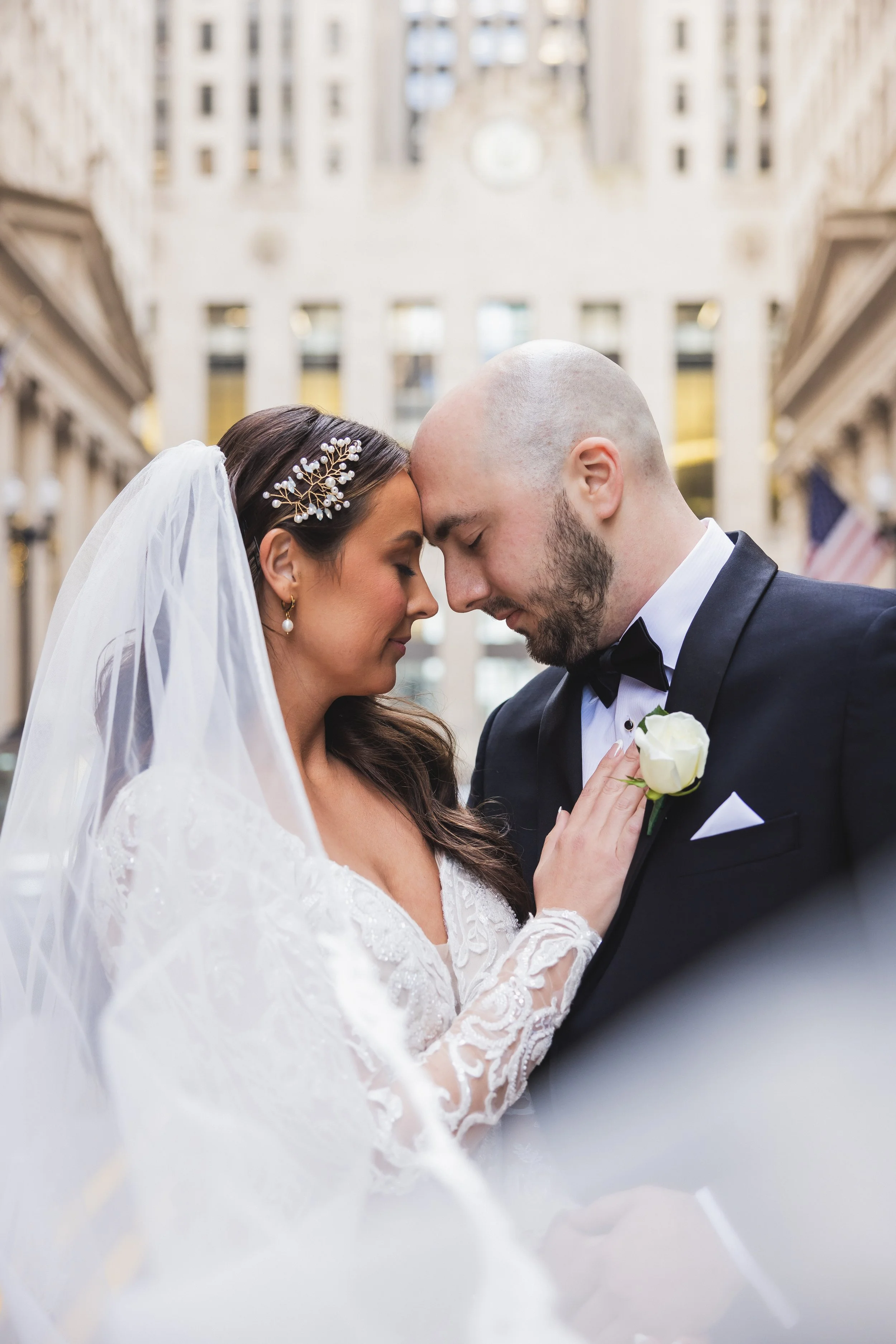 A bride and groom in wedding attire, gently touching foreheads, with an elegant building in the background. The bride wears a veil and lace dress, and the groom is in a tuxedo with a boutonniere.
