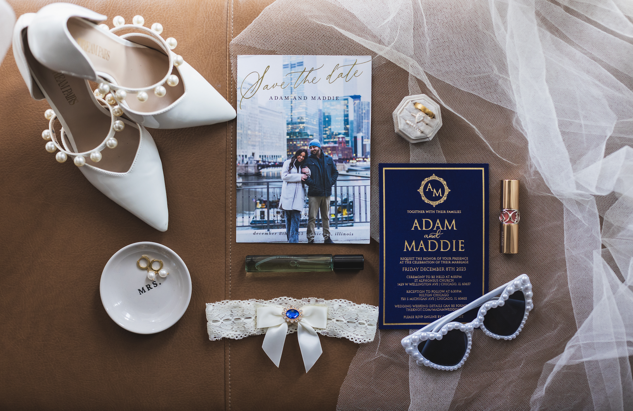 Wedding preparation items including pearl-embellished white shoes, a wedding invite, a save-the-date card, a ring on a fabric, sunglasses with pearls, lip balm, a lace garter, and a small dish labeled 'Mrs.' on a brown surface with a veil.