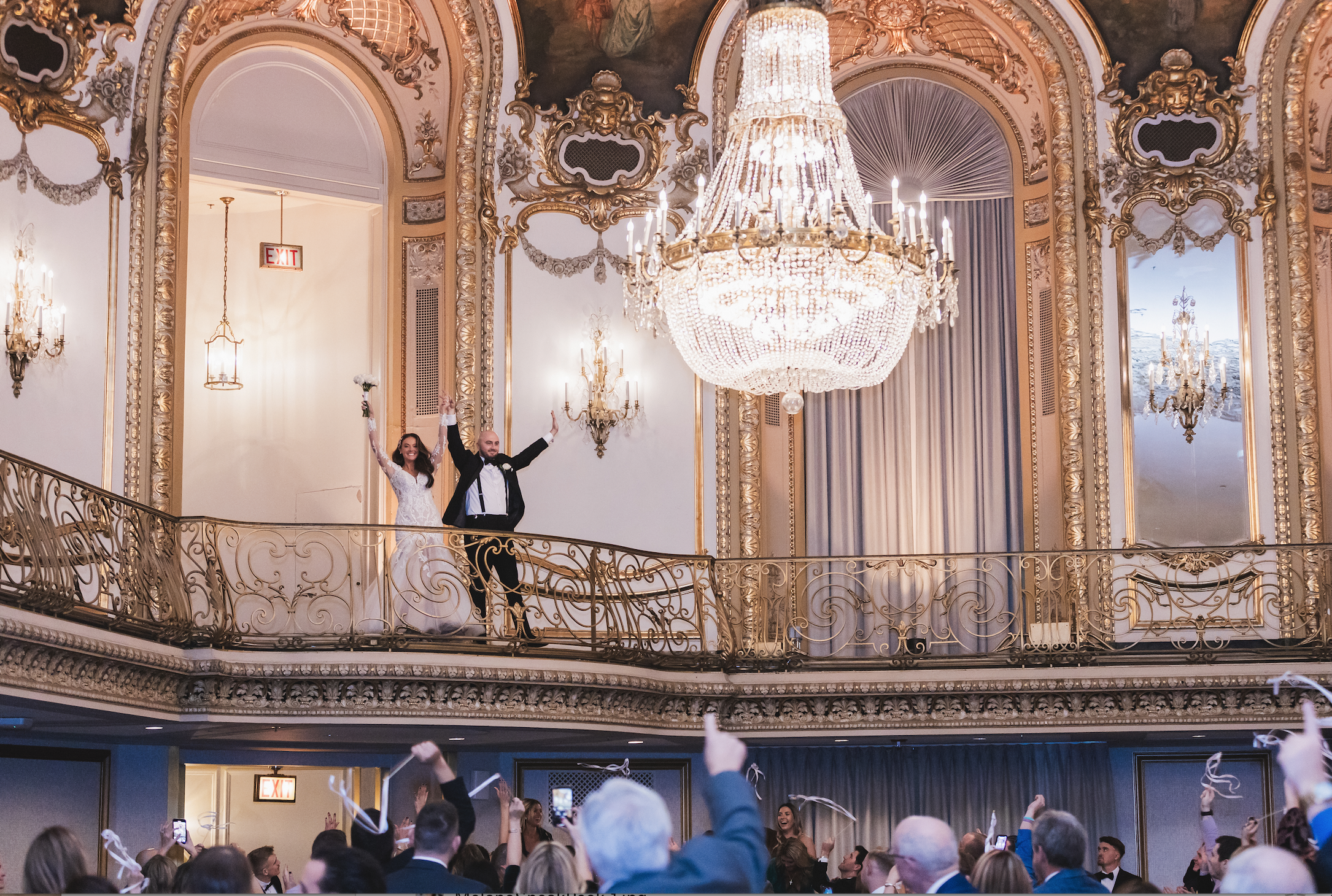A couple in wedding attire stands on a balcony waving to a cheering crowd in an elegant ballroom with ornate gold decor and a large chandelier.