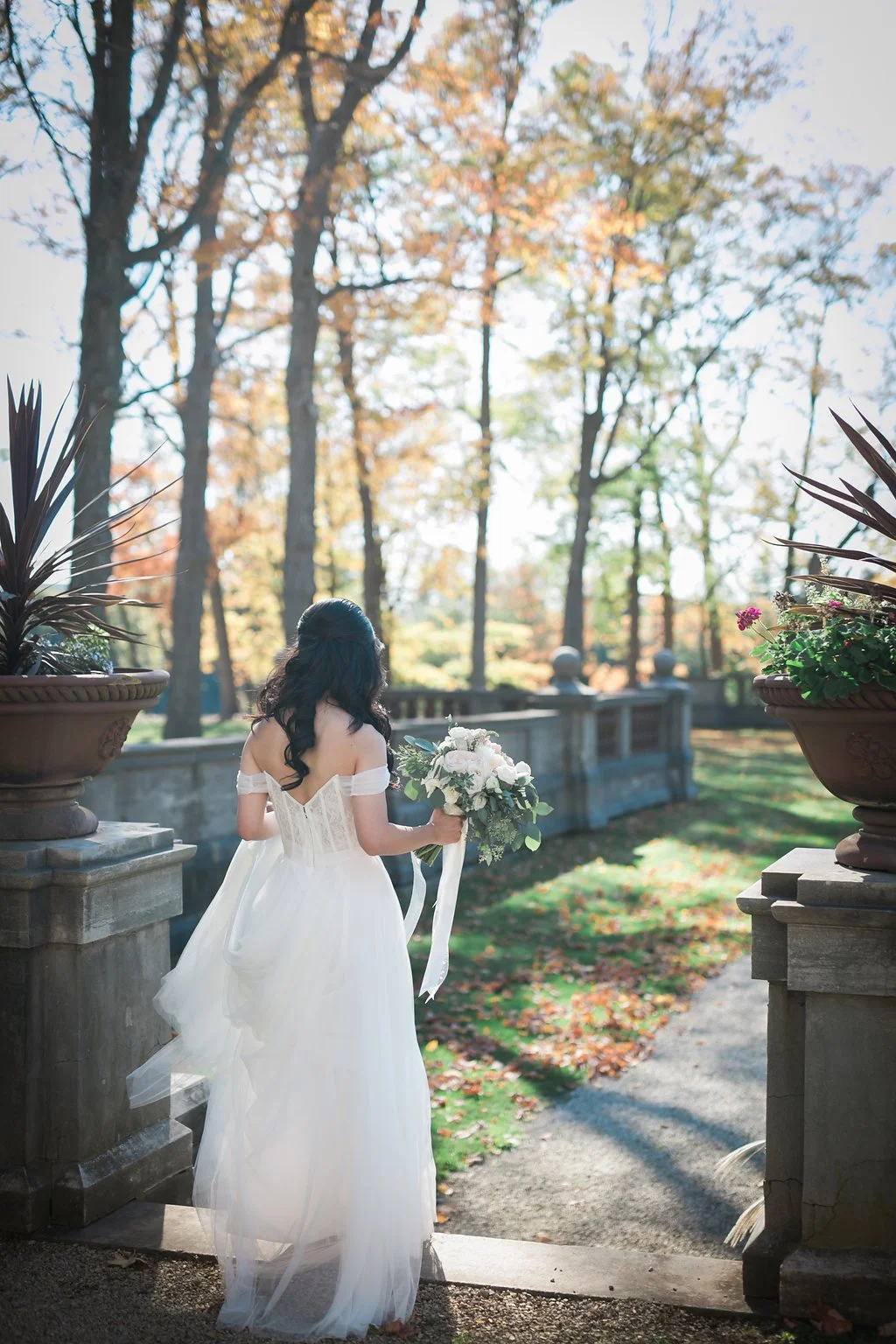 Bride in white dress holding bouquet standing outdoors among trees.