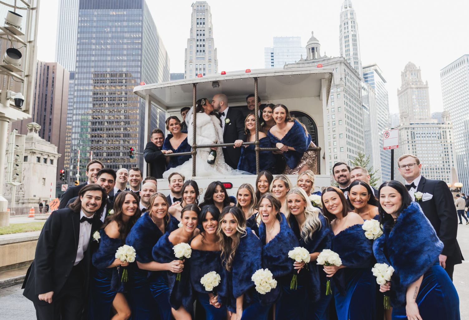 A large wedding party poses in front of a vintage streetcar in an urban setting. The group includes bridesmaids in blue outfits holding white bouquets and groomsmen in suits. The couple is kissing inside the streetcar. Skyscrapers and historic buildi