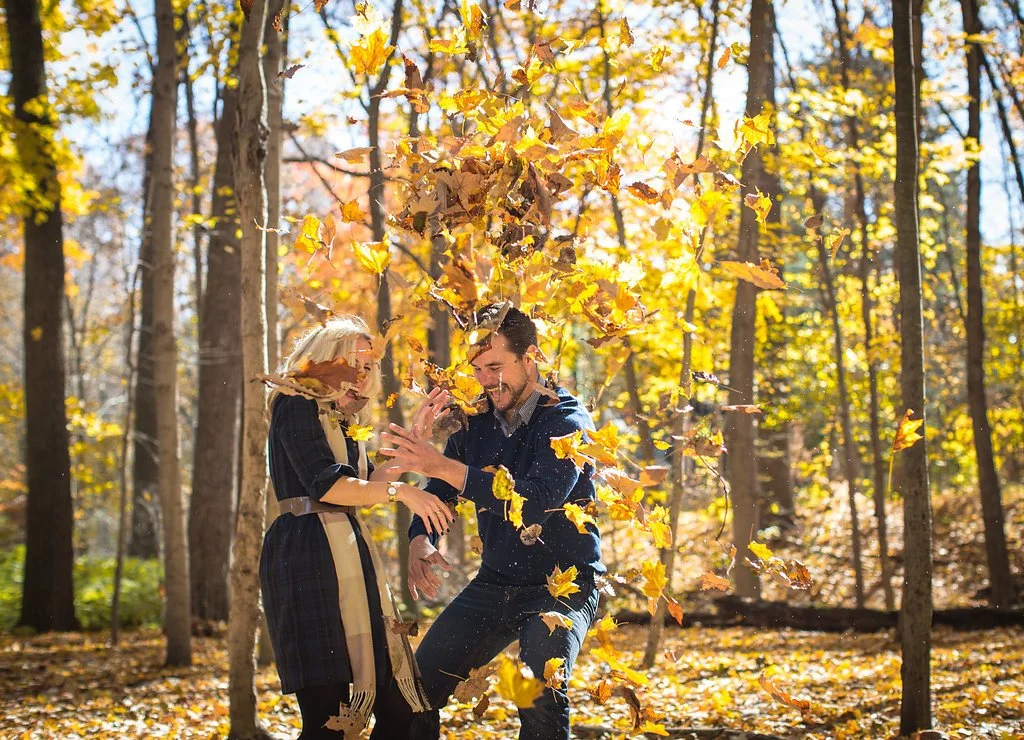 A man and woman playfully throwing autumn leaves in a sunny forest.