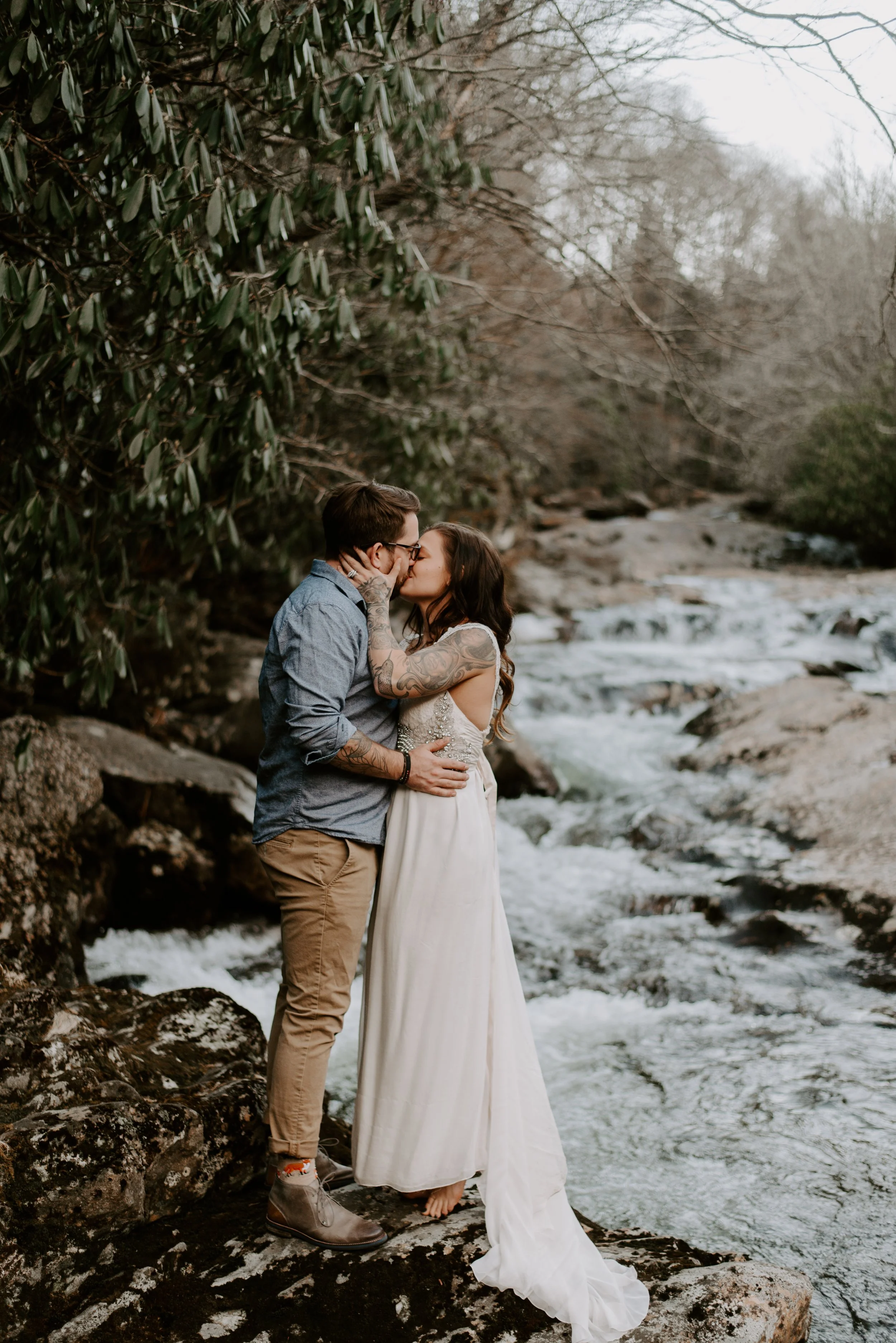 Couple kissing on rocky riverbank with greenery and stream in background.