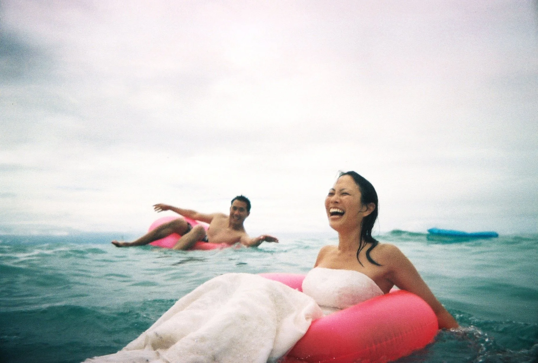 Two people in water on pink inflatable tubes, one wearing a white dress, both smiling and having fun in the ocean.