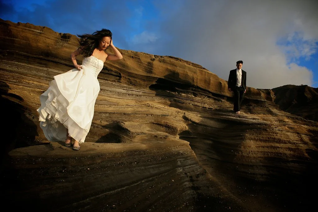 Bride in a white dress and groom in dark suit walking on rocky terrain under cloudy sky.